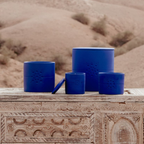 Bougies Izza Bleu de la marque Côté Bougie, senteur Mandarine Majorelle, exposées sur une table sculptée en bois, devant un paysage de désert marocain. Différents formats de bougies en céramique bleue sont mis en scène, pour une ambiance chaleureuse. Disponible chez Majorelle concept store marocain 