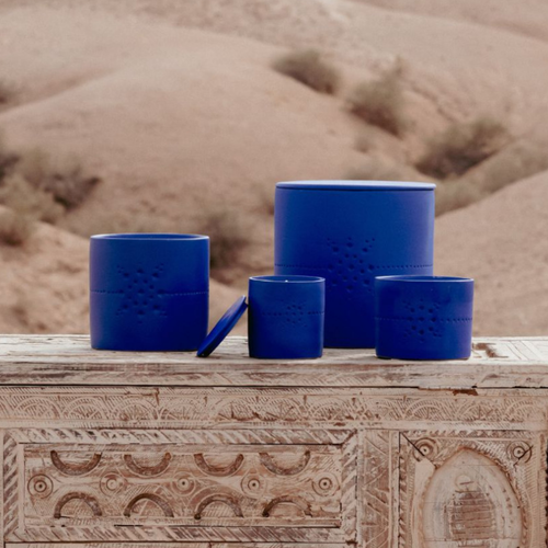 Bougies Izza Bleu de la marque Côté Bougie, senteur Mandarine Majorelle, exposées sur une table sculptée en bois, devant un paysage de désert marocain. Différents formats de bougies en céramique bleue sont mis en scène, pour une ambiance chaleureuse. Disponible chez Majorelle concept store marocain 
