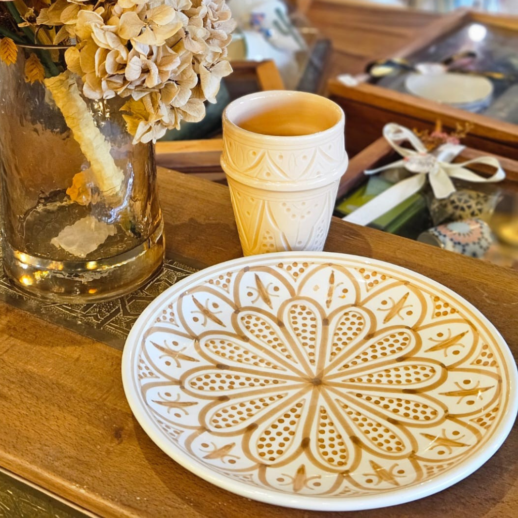 Assiette artisanale marocaine beldi, décor floral et géométrique couleur sable sur fond blanc, posée sur une table en bois, ambiance naturelle avec bouquet de fleurs séchées et tasse assortie.