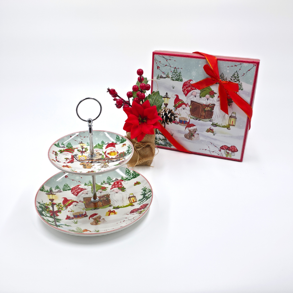 Présentoir à gâteaux de Noël à deux étages en porcelaine, décoré de motifs hivernaux avec lutins et sapins, présenté avec une boîte cadeau et une décoration florale rouge.