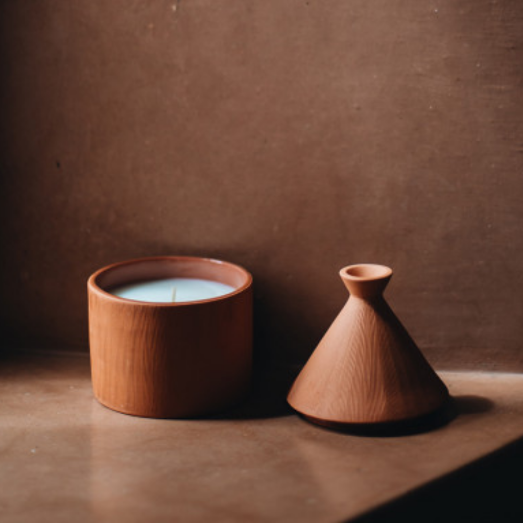 Bougie artisanale parfumée en forme de tajine en terre cuite terracotta, posée sur une étagère en bois brut. Son couvercle conique évoque les tajines marocains traditionnels. Un design élégant et épuré qui sublime la décoration intérieure avec une touche orientale chaleureuse.