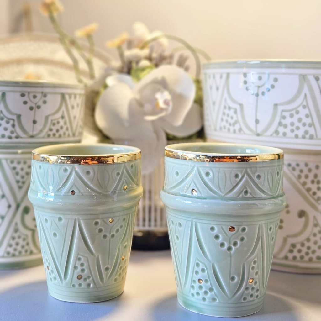 Tasse en céramique marocaine beldi, décor vert amande avec finitions dorées, posée en ambiance florale sur une table de service. Motifs ciselés à la main. Esprit raffiné et traditionnel.