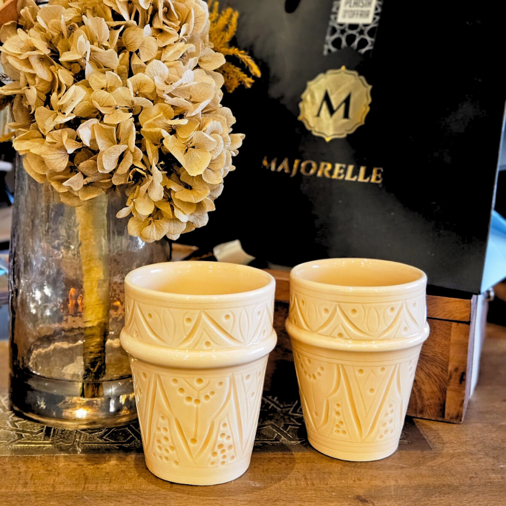 Tasse beldi artisanale beige sablé, décor marocain sculpté, posée sur une table bois ou métal, ambiance naturelle, bouquet de fleurs séchées à l’arrière-plan.