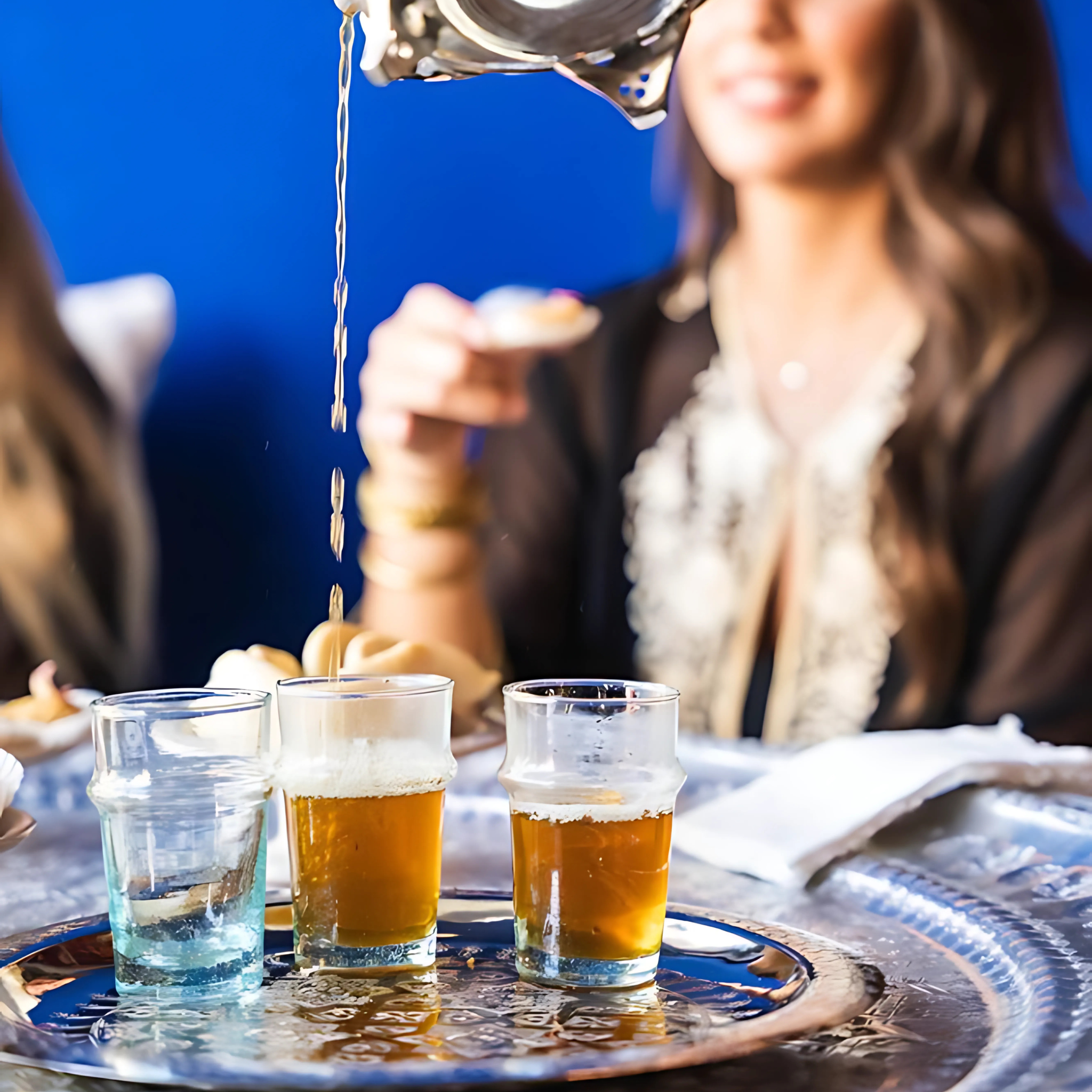 Photo de verres à thé marocains servis sur un plateau martelé, avec thé à la menthe en train d’être versé. Ambiance chaleureuse, femme en tenue traditionnelle au second plan, décor bleu Majorelle. Parfait pour illustrer l’art du thé marocain.