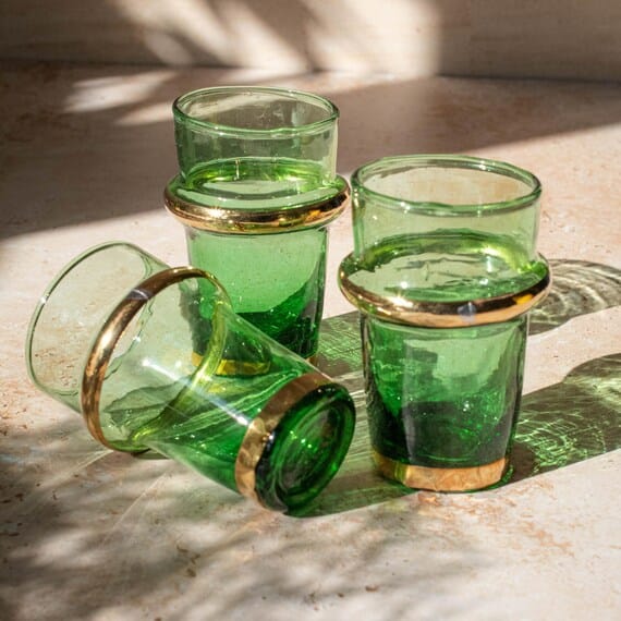 Verres à thé marocains verts avec double liseré doré, posés sur une table ensoleillée, ambiance artisanale et festive. Belles bulles dans le verre, lumière naturelle.